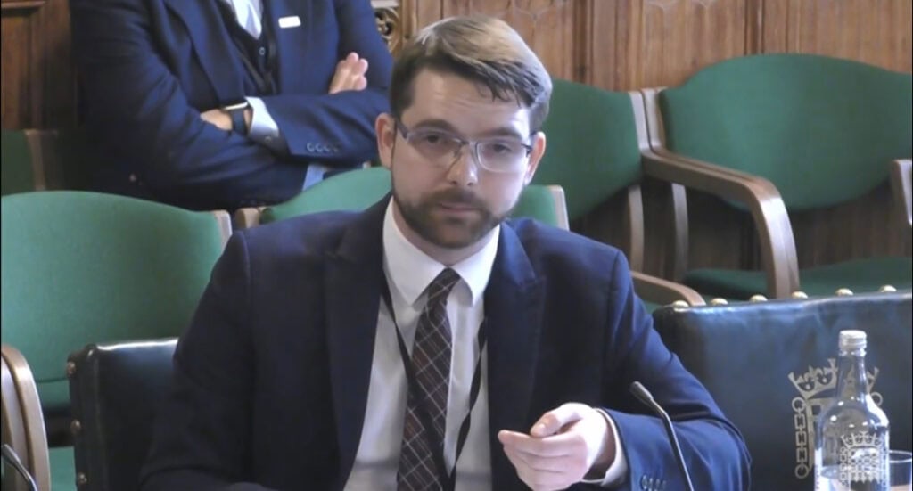 Evan wearing a suit sat at a table in parliament.  His hand is raised as he is making a point when he speaks.